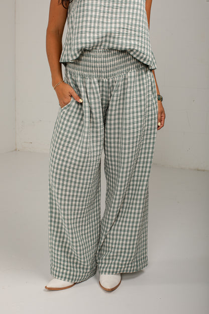 A person wearing wide-leg gingham pants in a muted sage green and white check pattern, paired with a matching top and white shoes, standing against a plain light gray wall.