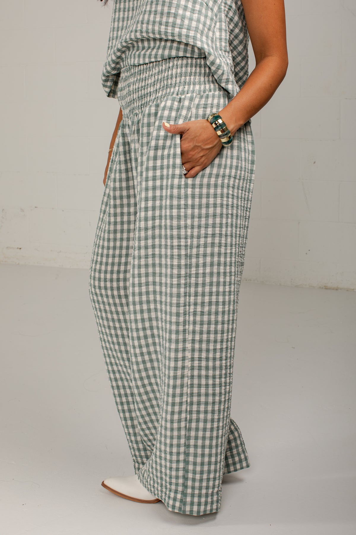A person wearing wide-leg gingham pants in a green and white check pattern, paired with a matching top and white pointed shoes, standing against a light gray studio wall.