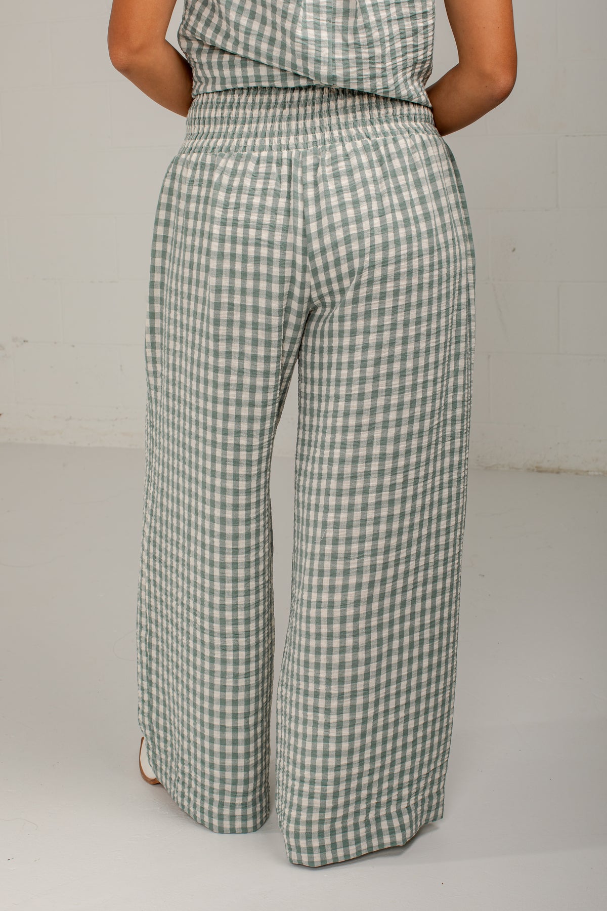 A person shown from the back wearing wide-leg cropped pants with a green and white gingham pattern, standing against a light gray wall.