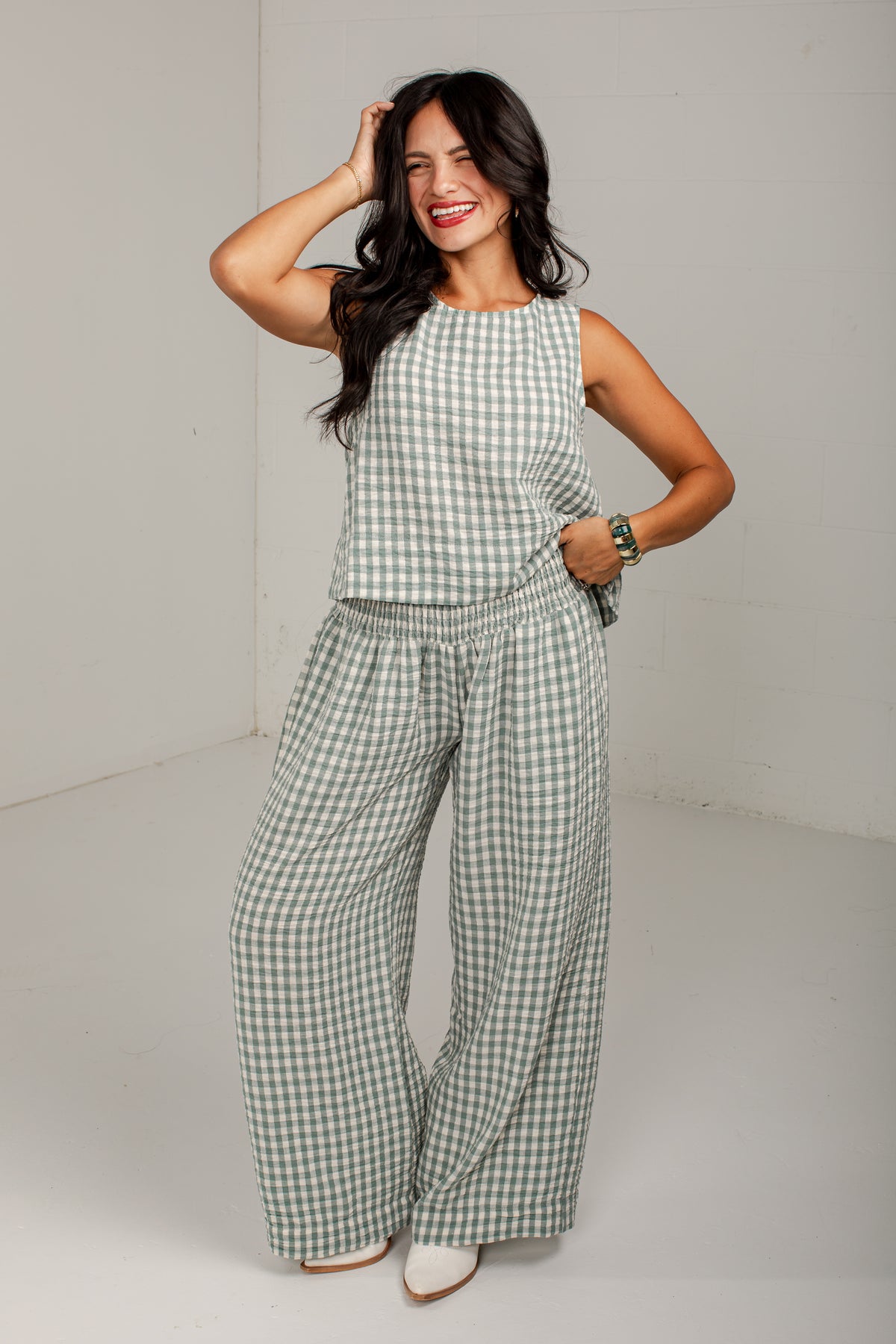 A woman modeling a sleeveless green and white gingham jumpsuit with wide-leg pants and an elastic waist, standing in a studio setting.