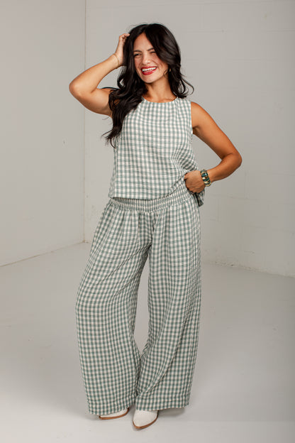 A woman modeling a sleeveless green and white gingham jumpsuit with wide-leg pants and an elastic waist, standing in a studio setting.