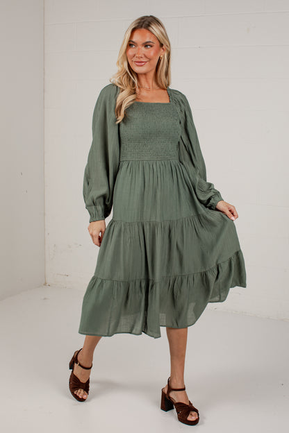 A woman modeling a sage green tiered midi dress with long balloon sleeves and a smocked bodice, paired with brown block-heel sandals, in a bright white studio setting.