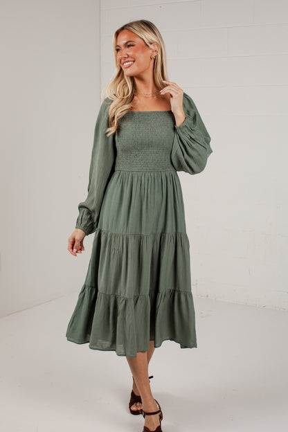 A woman modeling a sage green midi dress with long puff sleeves, a smocked bodice, and a tiered skirt, standing against a white brick wall background.