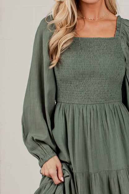 A woman wearing an olive green smocked dress with long balloon sleeves and a square neckline, shown from the shoulders to the hips against a light neutral background.