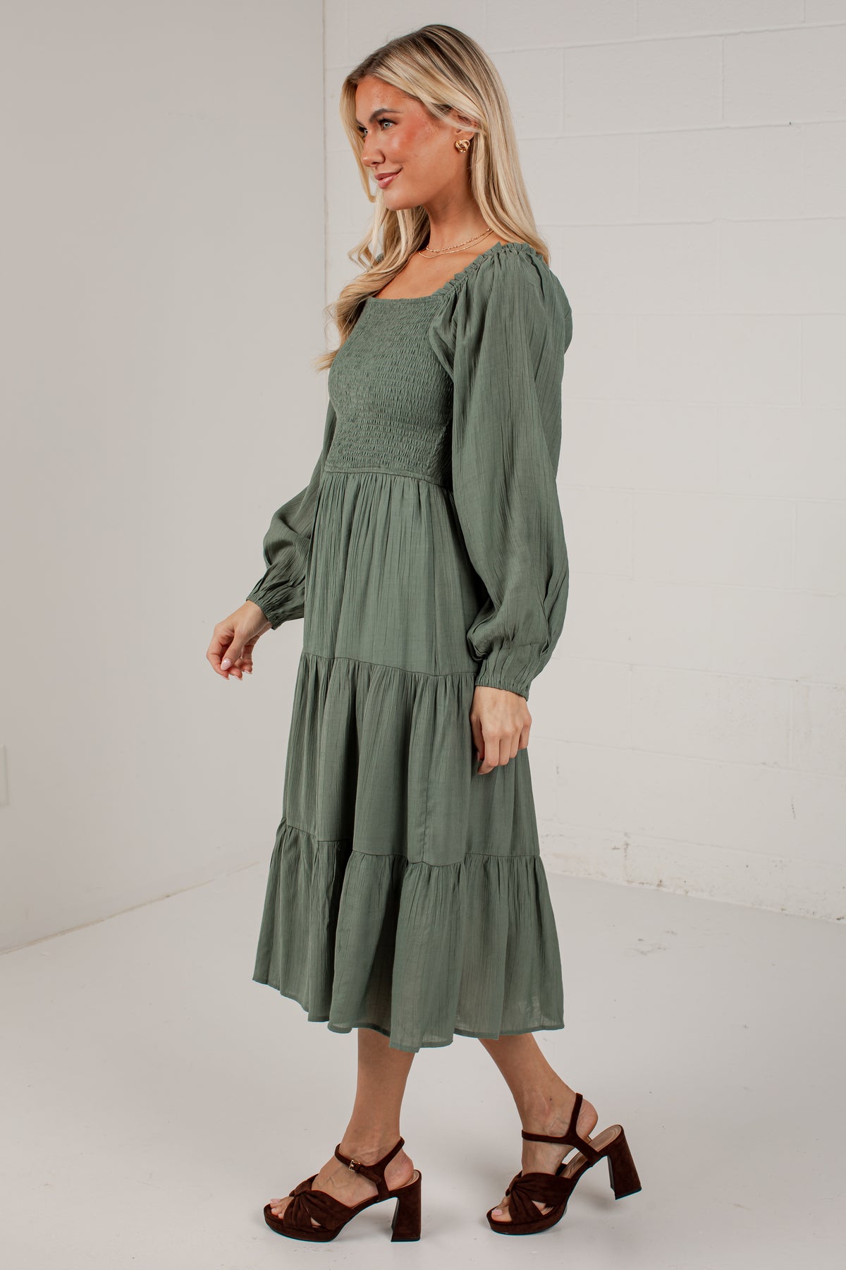 A woman modeling a sage green midi dress with long puff sleeves, a smocked bodice, and a tiered skirt, paired with dark brown heeled sandals against a white studio background.