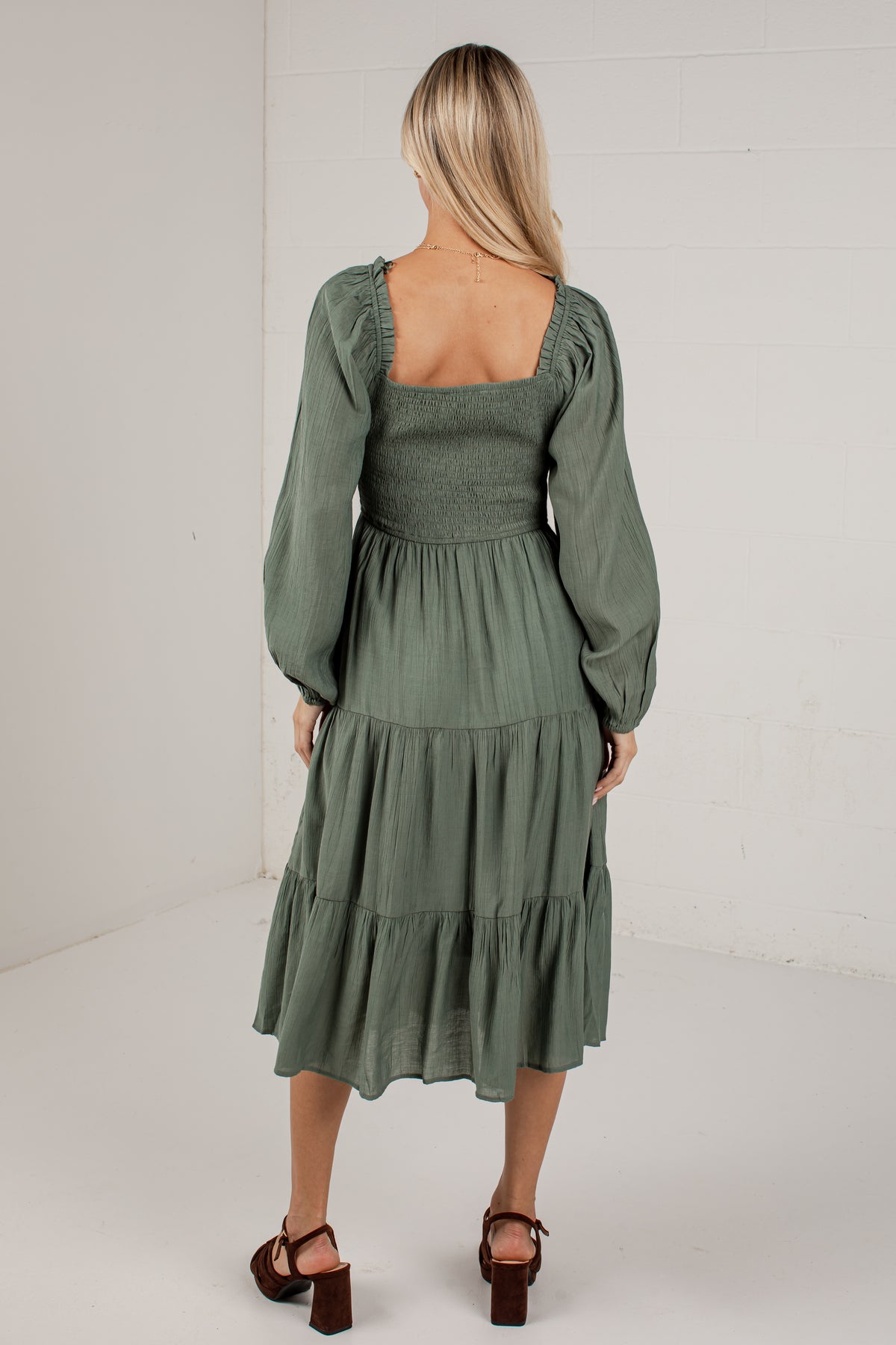 A woman viewed from behind wearing an olive green midi dress with long puff sleeves, a square neckline, and a tiered skirt, paired with brown block-heel sandals in a bright studio setting.