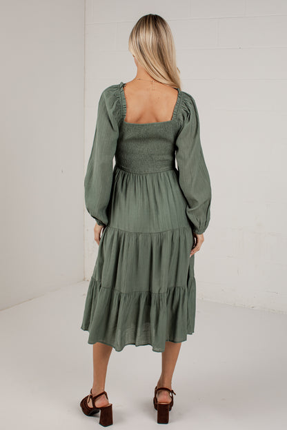 A woman viewed from behind wearing an olive green midi dress with long puff sleeves, a square neckline, and a tiered skirt, paired with brown block-heel sandals in a bright studio setting.
