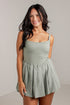 A woman modeling a sage green sleeveless mini dress with a fitted bodice and flared pleated skirt, standing against a light gray studio wall.