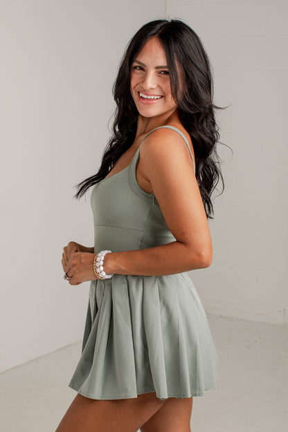 A woman modeling a sage green sleeveless skater dress with a pleated skirt, standing against a plain light gray studio background.