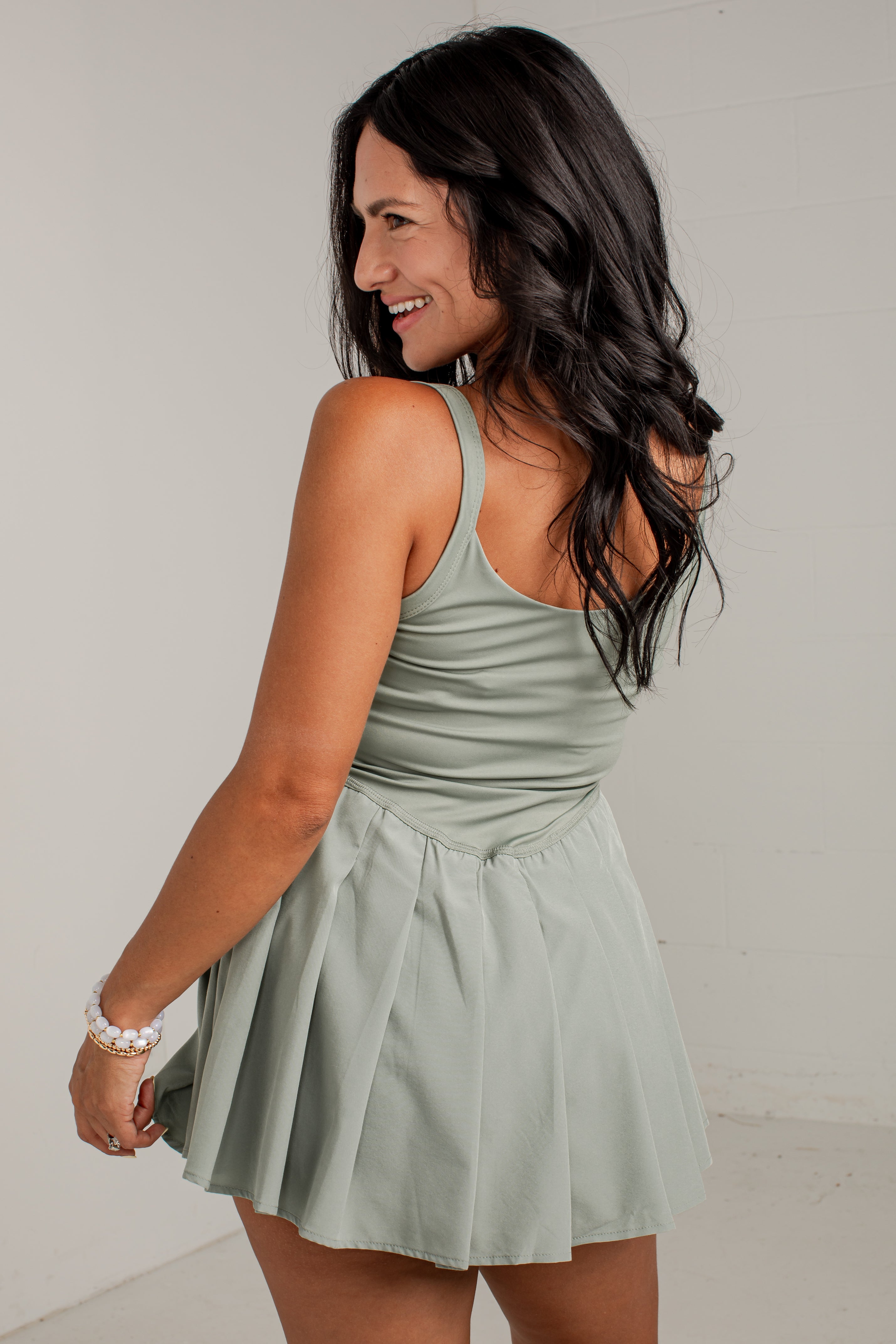 A woman with long dark hair is shown from the back wearing a sage green sleeveless mini dress with a pleated skirt, set against a plain light gray background.