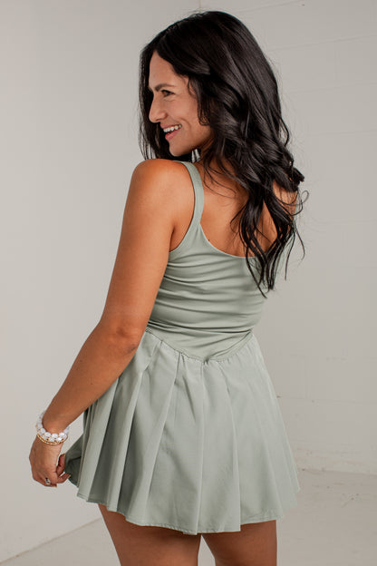 A woman with long dark hair is shown from the back wearing a sage green sleeveless mini dress with a pleated skirt, set against a plain light gray background.