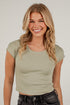 A smiling blonde woman wearing a ribbed sage green short-sleeve crop top and dark gray jeans, standing against a white brick wall.