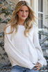A woman wearing a chunky white knit sweater and light blue jeans, standing in front of decorated Christmas trees with warm lights.