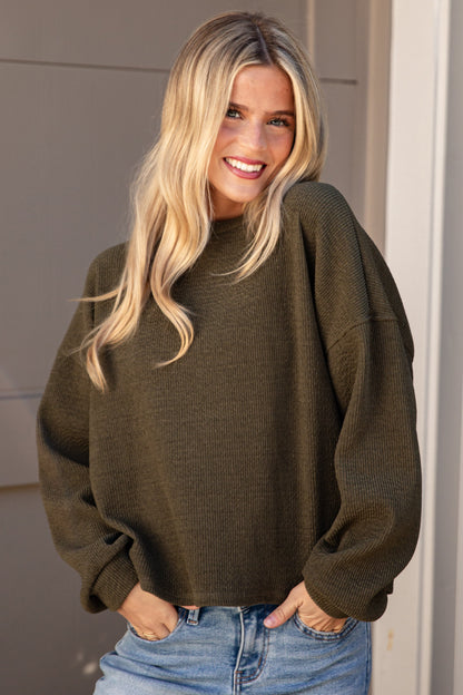 A smiling blonde woman wearing an oversized olive green knit sweater and light blue jeans, standing in front of a beige garage door and white brick wall.