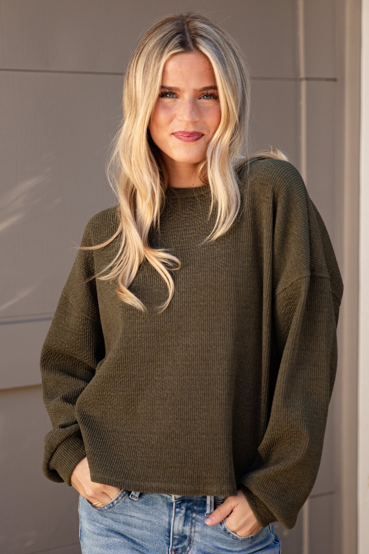 A blonde woman wearing an oversized olive green knit sweater and light blue jeans, standing in front of a neutral-toned paneled wall.