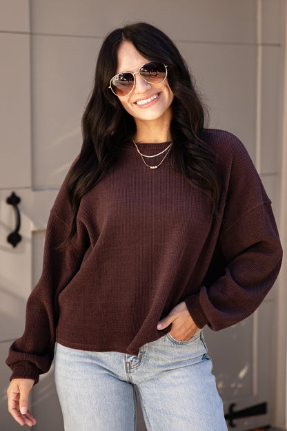 A woman wearing a dark brown waffle-knit long-sleeve sweater and light blue jeans, standing in front of a modern paneled wall.