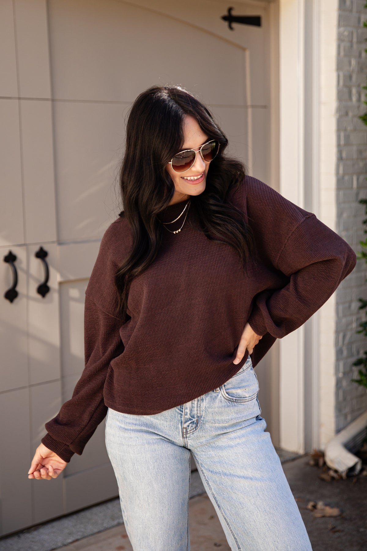 A woman wearing a dark brown long-sleeve crewneck sweatshirt and light wash blue jeans, standing in front of a beige garage door.