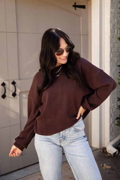 A woman wearing a dark brown long-sleeve crewneck sweatshirt and light wash blue jeans, standing in front of a beige garage door.