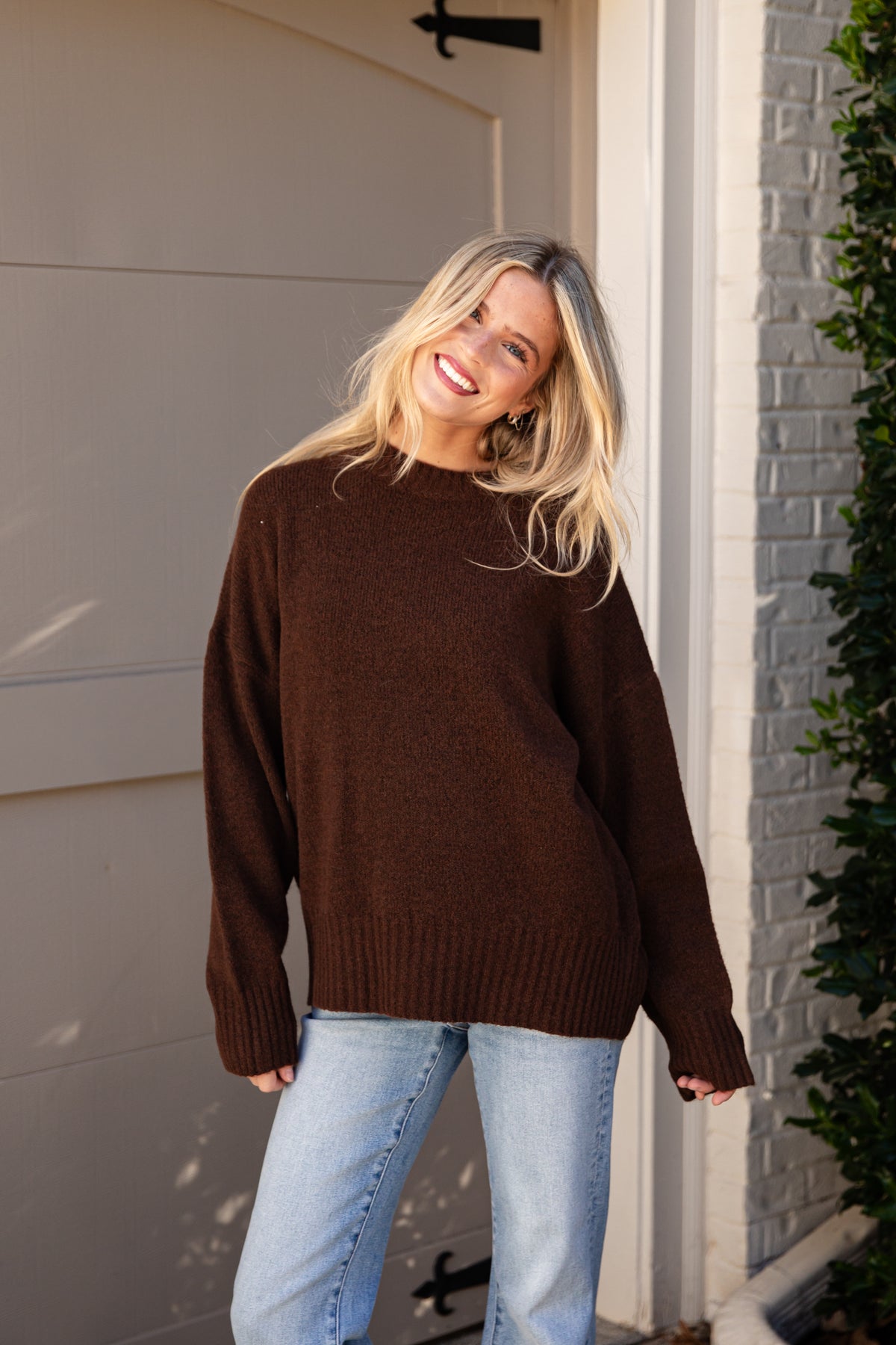 A woman wearing an oversized dark brown knit sweater and light blue jeans, standing outdoors in front of a garage door and brick wall.