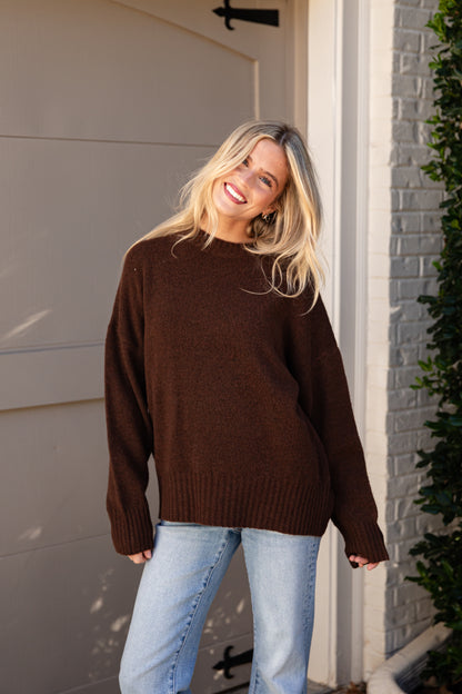 A woman wearing an oversized dark brown knit sweater and light blue jeans, standing outdoors in front of a garage door and brick wall.