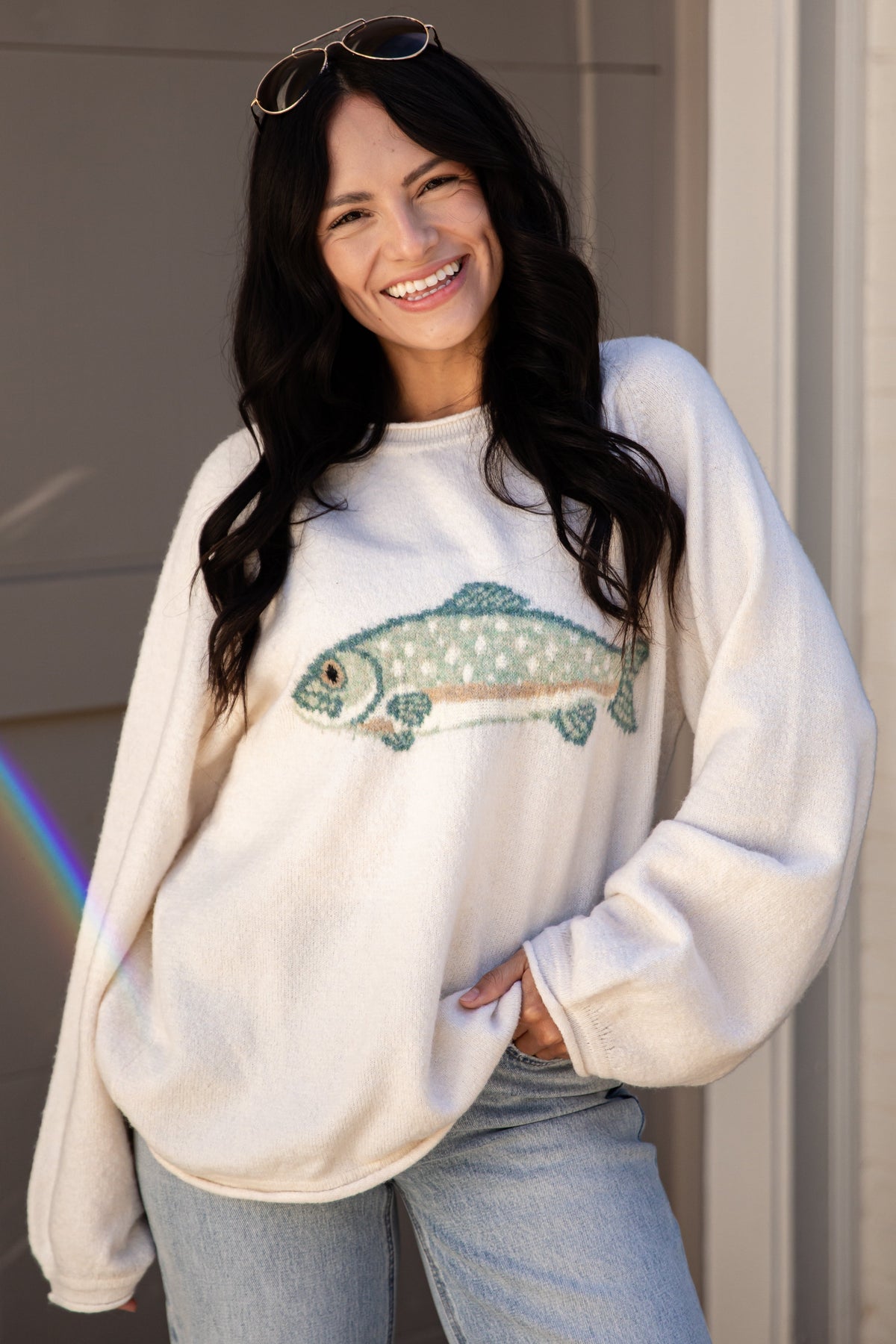 A woman smiling while wearing a cream-colored crewneck sweatshirt with a detailed fish graphic on the front, paired with light blue jeans and sunglasses on her head.