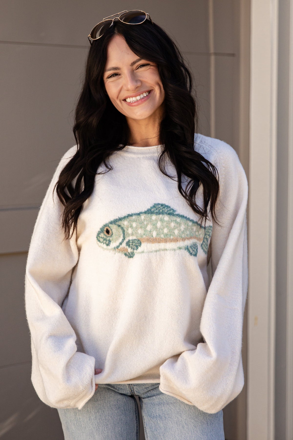 A woman with long dark hair and sunglasses on her head is smiling while wearing a cream-colored crewneck sweatshirt with a fish graphic on the front, paired with light blue jeans.