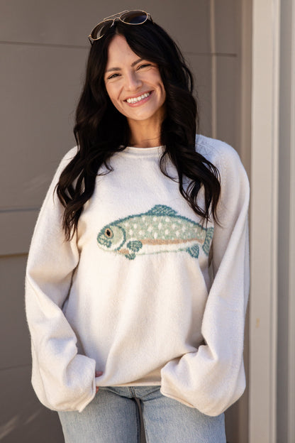 A woman with long dark hair and sunglasses on her head is smiling while wearing a cream-colored crewneck sweatshirt with a fish graphic on the front, paired with light blue jeans.