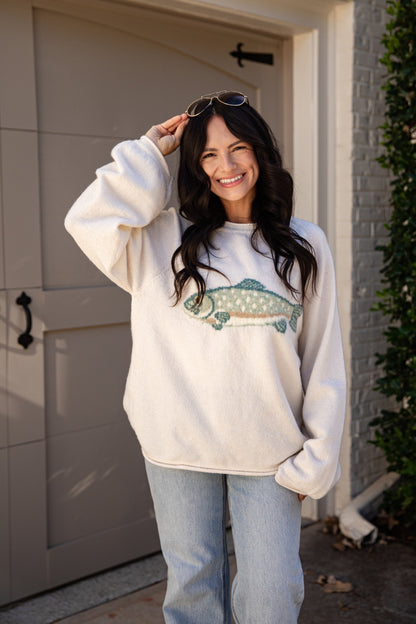 A woman wearing a cream-colored fleece sweatshirt with a green and white fish graphic on the front, paired with light blue jeans, standing in front of a garage door.