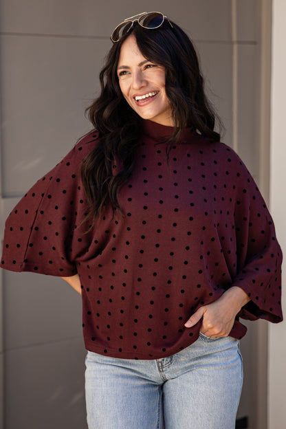 A woman wearing a burgundy mock-neck top with black polka dots and light-wash blue jeans, standing in front of a beige paneled wall.