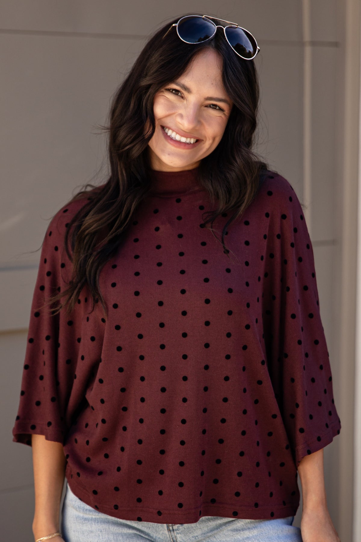 A woman wearing a burgundy mock-neck top with black polka dots and light-wash blue jeans, standing outdoors in front of a beige door. She has sunglasses on her head and a bracelet on her wrist.