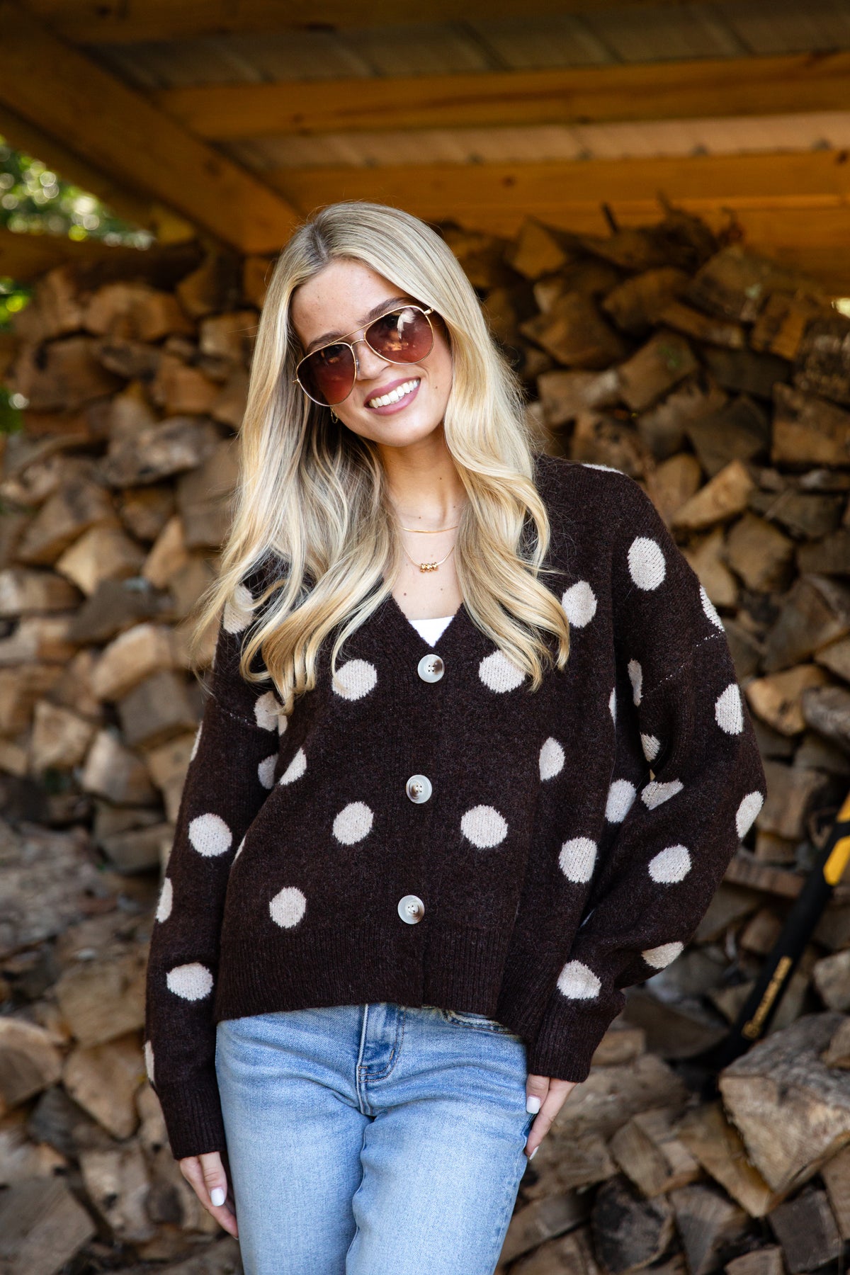 A woman with blonde hair and sunglasses wearing a dark brown cardigan with white polka dots and light blue jeans, standing in front of a stacked woodpile.