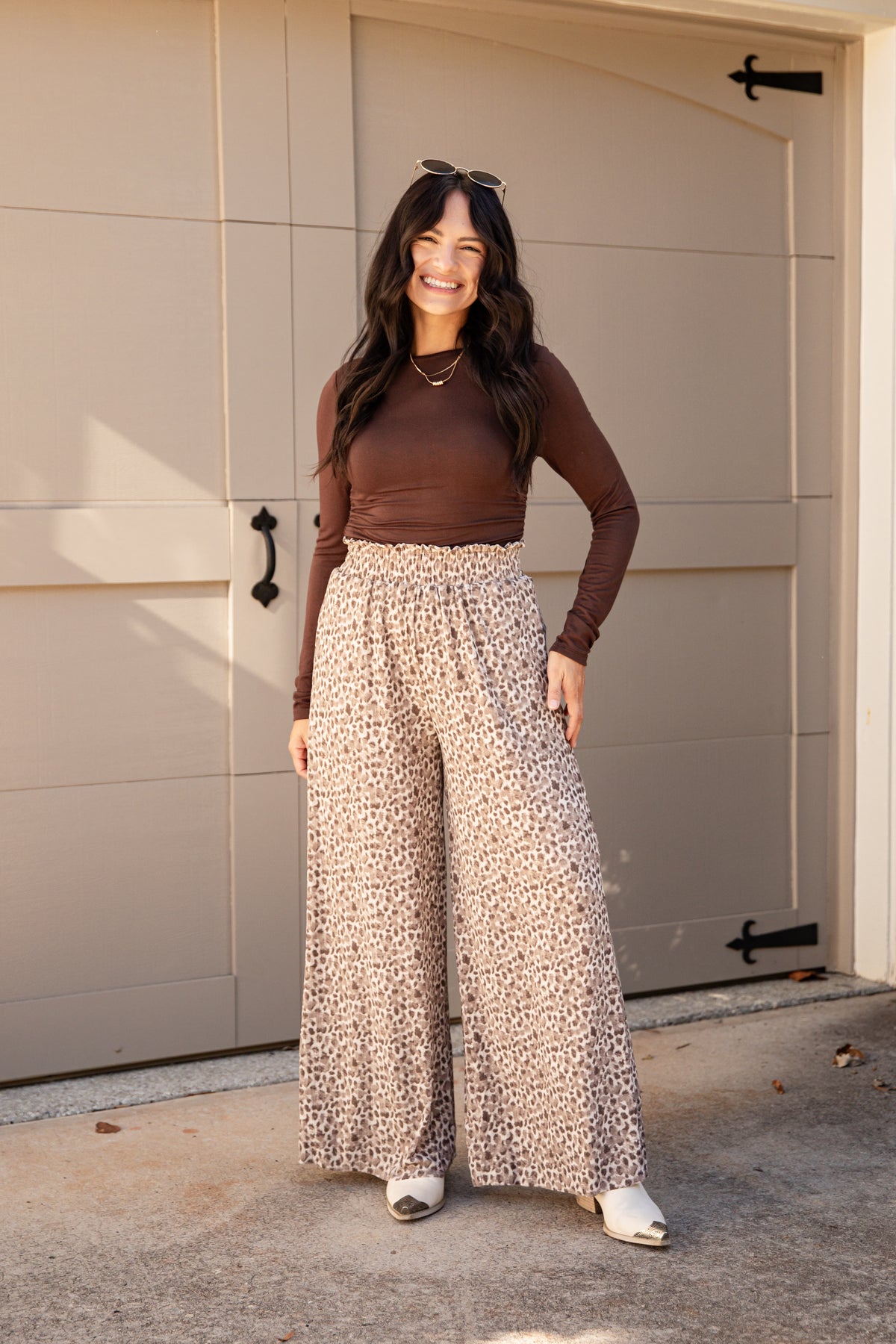 A woman stands in front of a beige garage door wearing a brown long-sleeve top and high-waisted, wide-leg beige pants with a small dark print. She is smiling and wearing white sneakers.