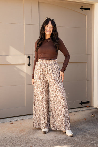 A woman stands in front of a beige garage door wearing a brown long-sleeve top and high-waisted, wide-leg beige pants with a small dark print. She is smiling and wearing white sneakers.
