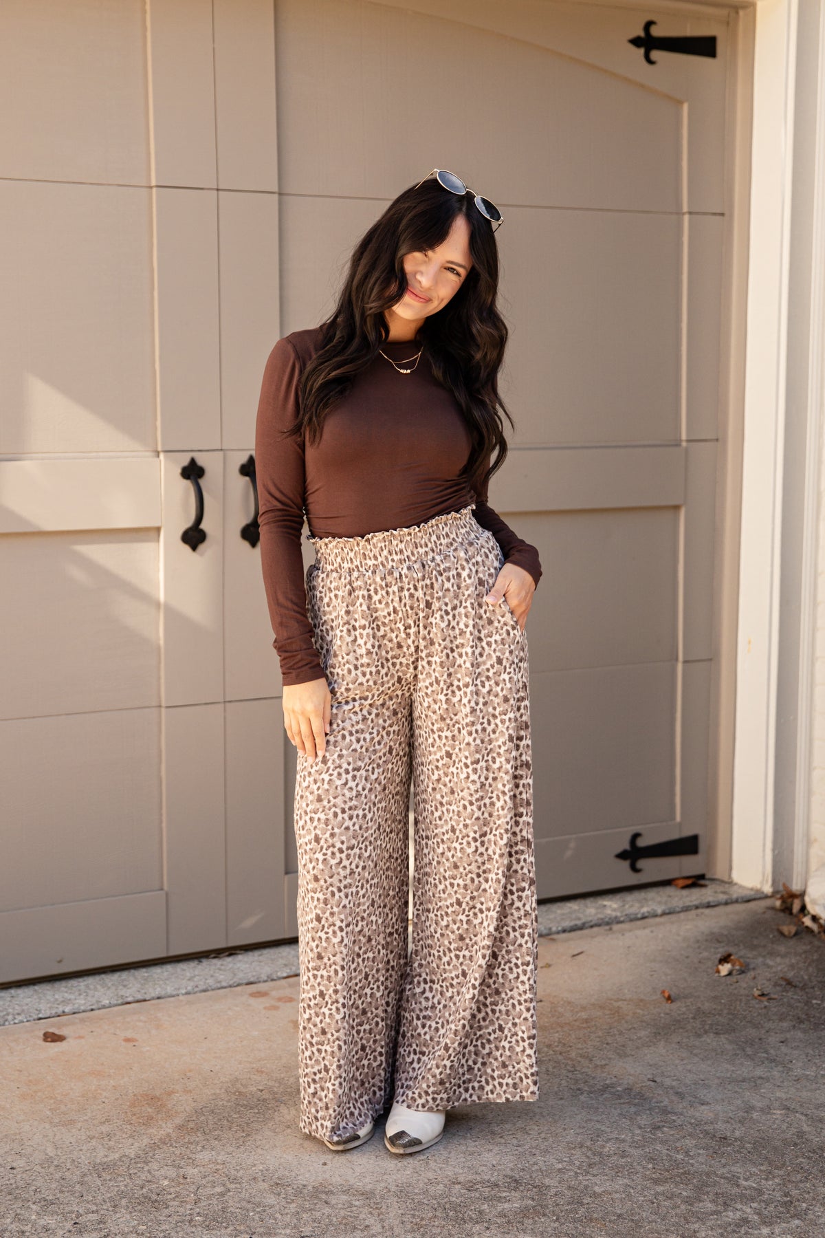 A woman stands in front of a beige garage door wearing a dark brown long-sleeve top and high-waisted, wide-leg pants with a beige, brown, and black leopard print. She is wearing white slide sandals and sunglasses are resting on her head.