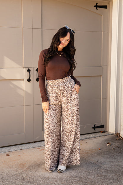 A woman stands in front of a beige garage door wearing a dark brown long-sleeve top and high-waisted, wide-leg pants with a beige, brown, and black leopard print. She is wearing white slide sandals and sunglasses are resting on her head.