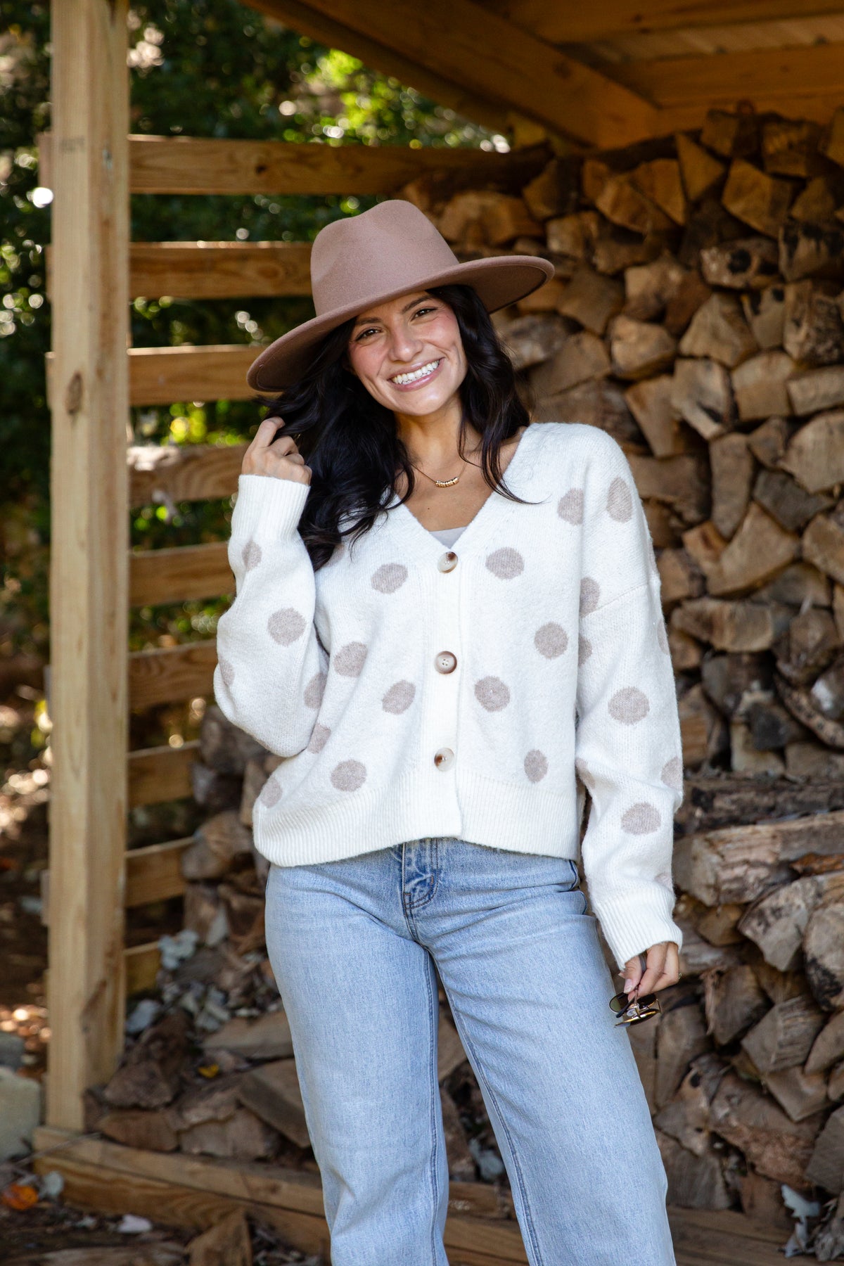 A woman wearing a cream knit cardigan with brown polka dots, a brown wide-brim hat, and light blue jeans, standing in front of a rustic wood pile.