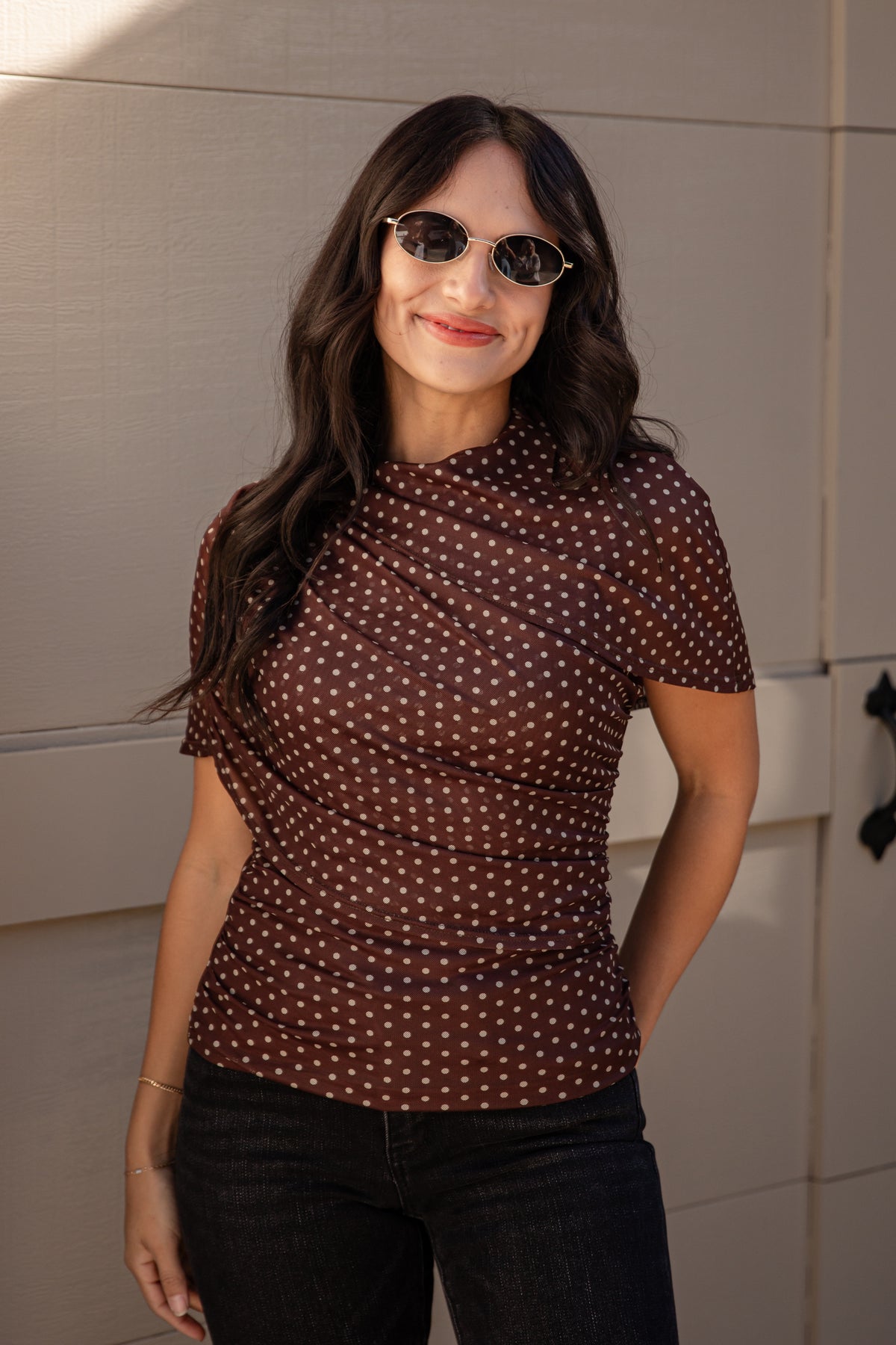 A woman wearing a brown short-sleeve top with a white polka dot pattern, styled with dark jeans and sunglasses, standing against a beige paneled wall.