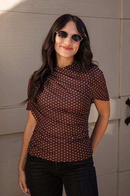 A woman wearing a brown short-sleeve top with a white polka dot pattern, styled with dark jeans and sunglasses, standing against a beige paneled wall.