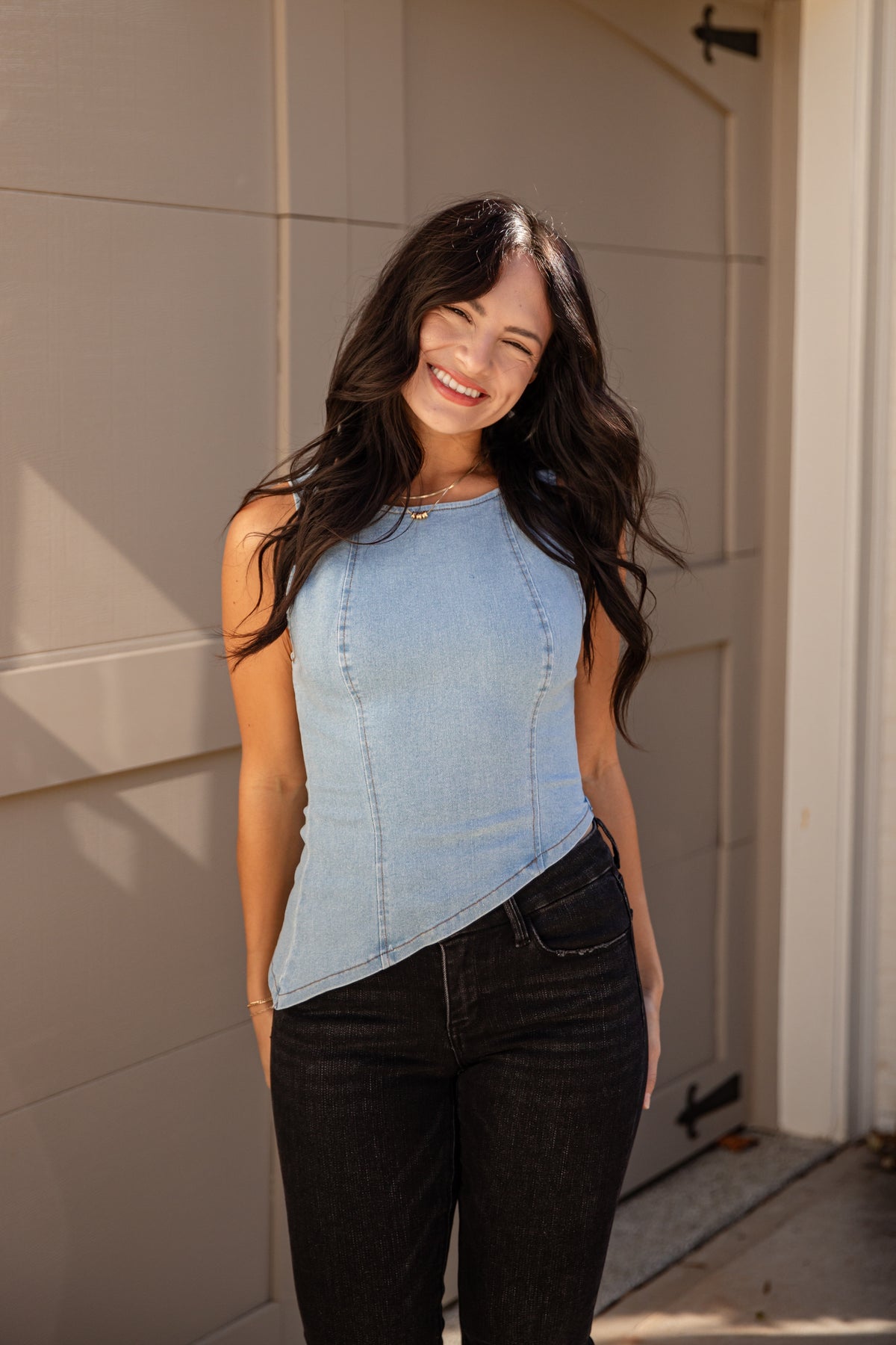 A woman wearing a light blue sleeveless asymmetrical top and black jeans, standing in front of a beige garage door.