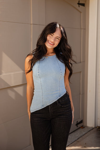 A woman wearing a light blue sleeveless asymmetrical top and black jeans, standing in front of a beige garage door.