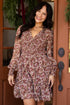 A woman stands in a doorway wearing a long-sleeve floral mini dress with a ruffled V-neck and a matching waist tie. The dress has a dense pattern of pink, red, and green flowers on a dusty mauve background.