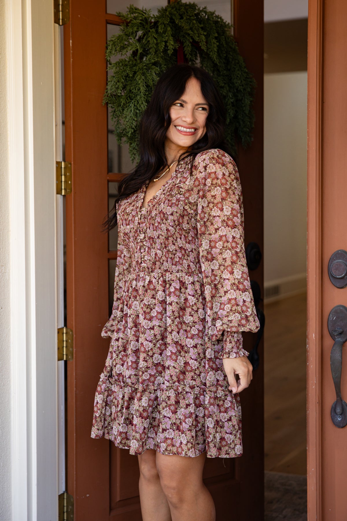 A woman wearing a long-sleeve floral mini dress with a ruffled hem, standing in a doorway with a green wreath on the door behind her.