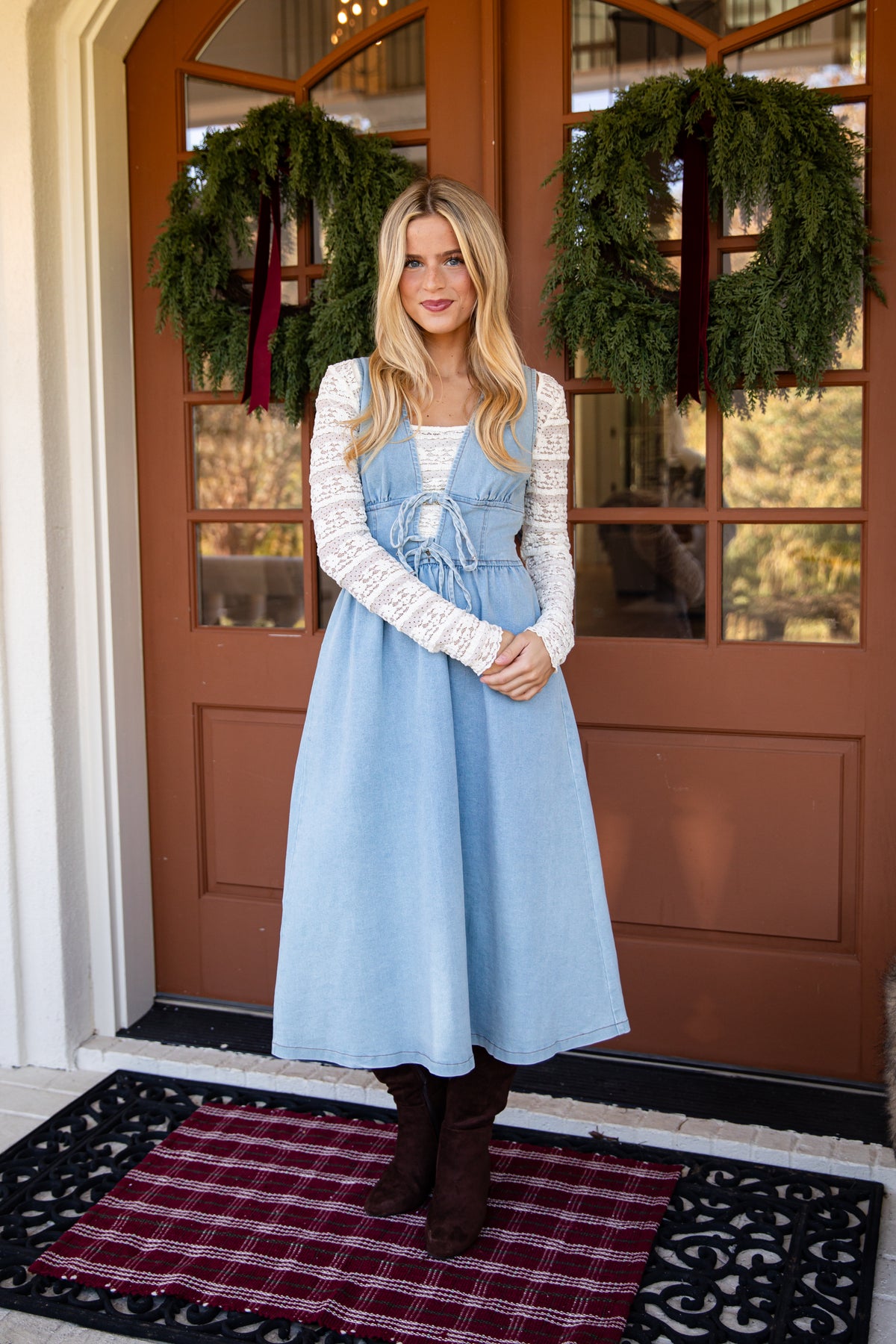 A woman stands on a front porch wearing a light blue denim pinafore dress with a white lace long-sleeve top layered underneath, paired with dark brown boots. The background features a brown wooden door with green holiday wreaths.