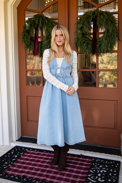 A woman stands on a front porch wearing a light blue denim pinafore dress with a white lace long-sleeve top layered underneath, paired with dark brown boots. The background features a brown wooden door with green holiday wreaths.