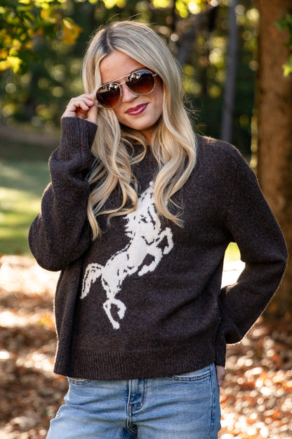 A woman wearing a dark brown knit sweater with a white horse graphic and light blue jeans, standing outdoors in a park with trees and fallen leaves in the background.
