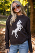 A woman wearing a dark brown knit sweater with a white horse graphic, paired with blue jeans and sunglasses, standing outdoors in a park with trees and fallen leaves.