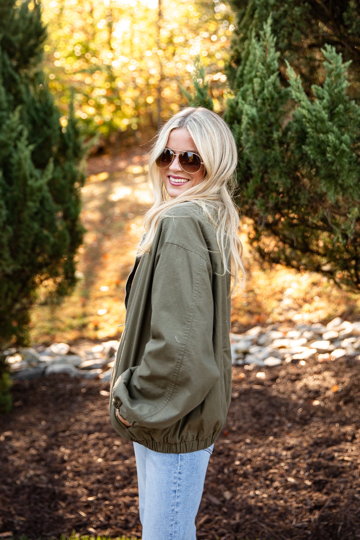 A woman with blonde hair and sunglasses wearing an olive green jacket and light blue jeans, standing outdoors in a wooded setting with trees and mulch.