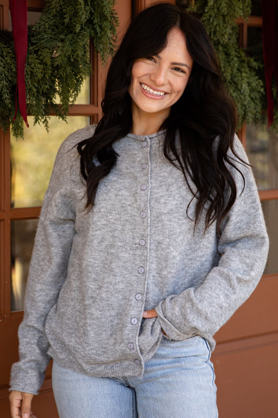 A woman smiling and wearing a light gray heathered button-front cardigan sweater and light blue jeans, standing in front of a warm brown door decorated with green garlands.