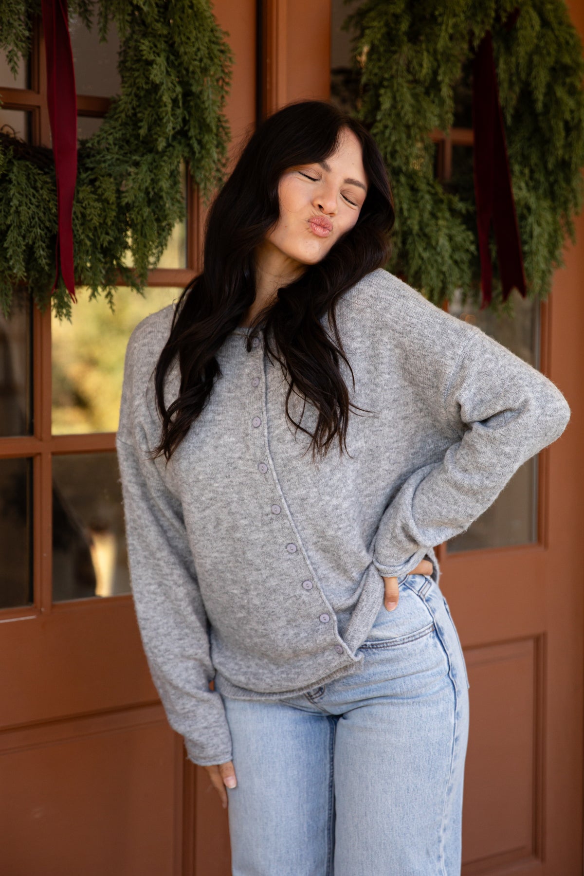 A woman wearing a light gray heathered knit cardigan and light blue jeans, posing in front of a rustic brown wooden door decorated with green holiday wreaths.