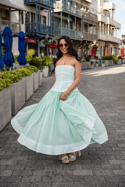 Woman in a light green strapless dress standing on a street with buildings and plants in the background.